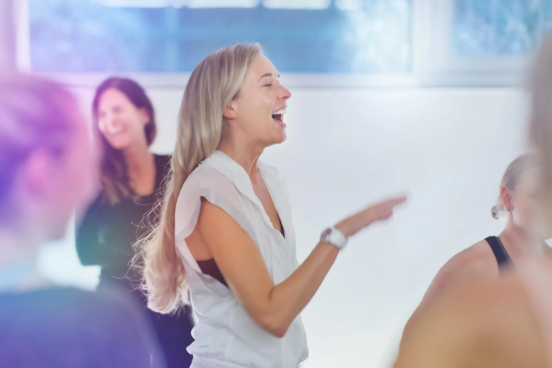 Suzanne lacht in weissem Top, zeigt mit dem Finger, Gruppe im Hintergrund